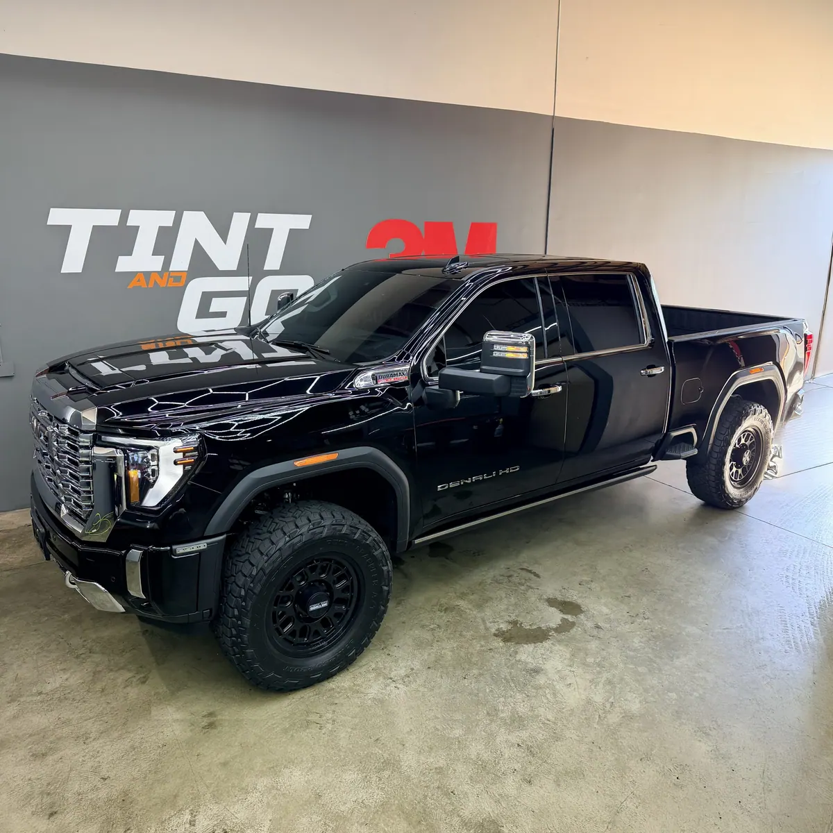 Black GMC Sierra Denali heavy-duty pickup with a chrome grille and tinted glass under the shop's wall graphic