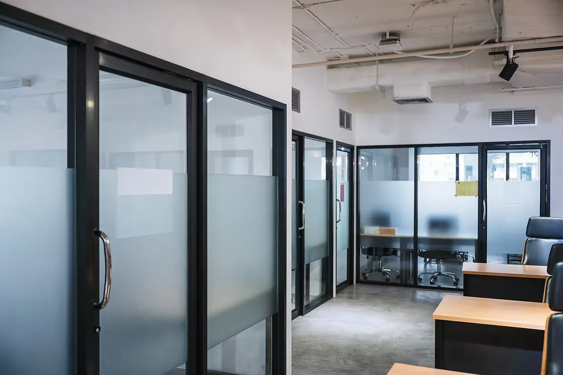 Office interior where glass partition walls and doors carry frosted privacy bands, desks and a meeting room beyond