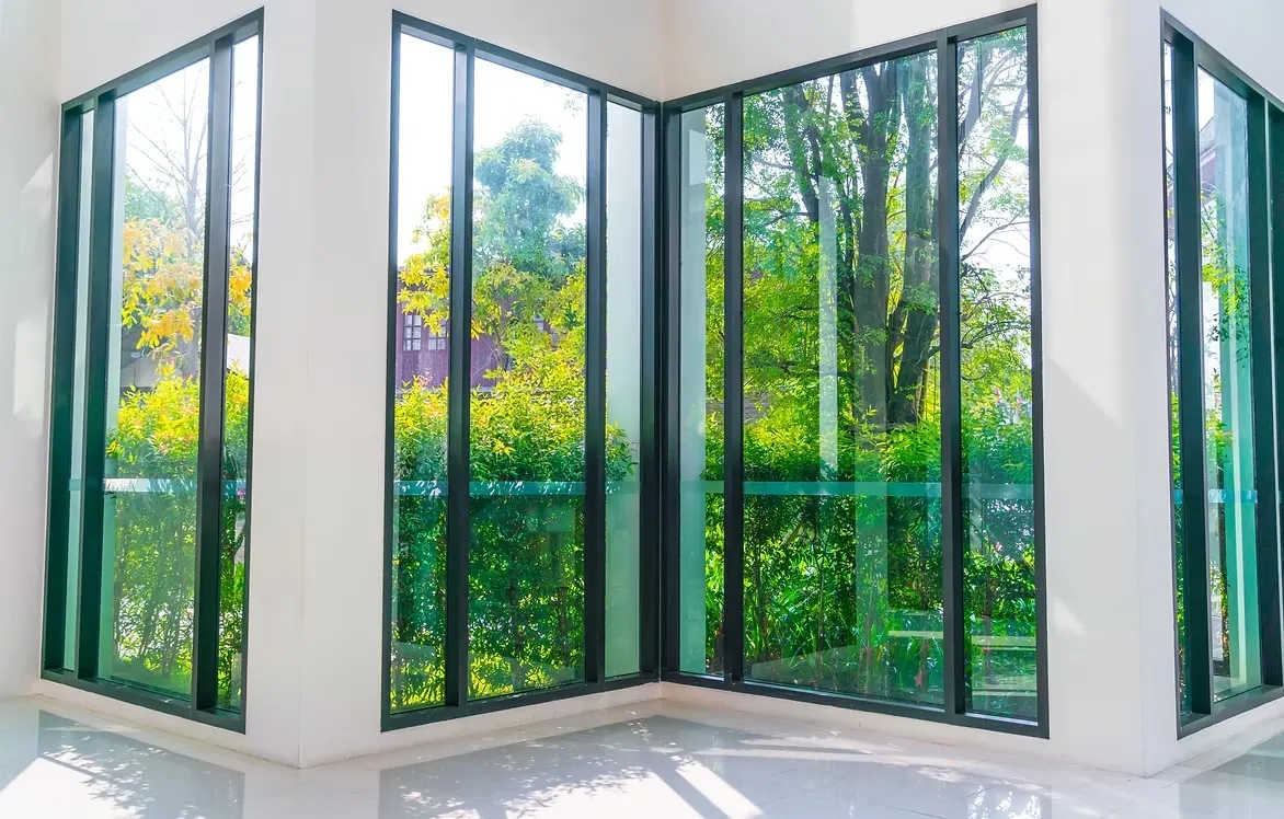 Sunlit corner of a home with floor-to-ceiling black-framed windows looking out on trees and garden planting