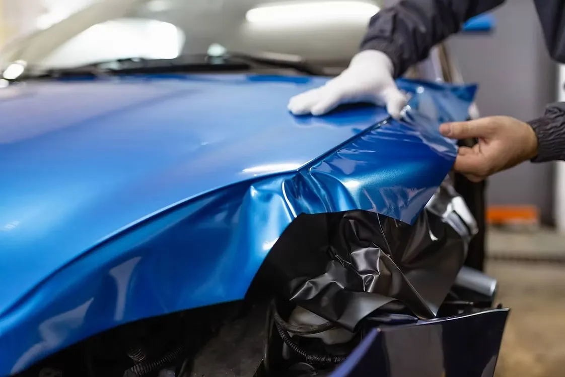 Gloved installer laying gloss blue vinyl wrap over a car's front fender as the backing liner peels away